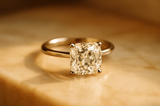 A single cushion cut diamond solitaire engagement ring with a soft pillow shape and large open facets, on a slim gold band resting on warm marble.