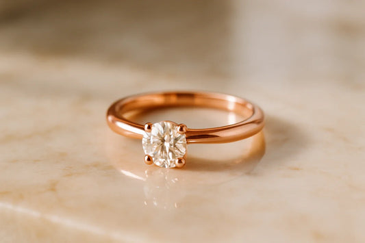 A single rose gold solitaire engagement ring with one round diamond in a four-prong setting, the warm pink-gold band resting on cream marble.