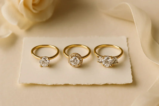 Three engagement rings side-by-side on cream paper: solitaire on the left, halo in the middle, and three-stone on the right, all in yellow gold.